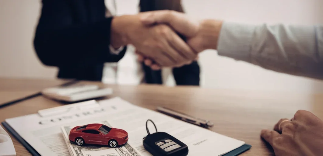 Two people shaking hands over a desk holding a signed contract, car keys and a red toy car