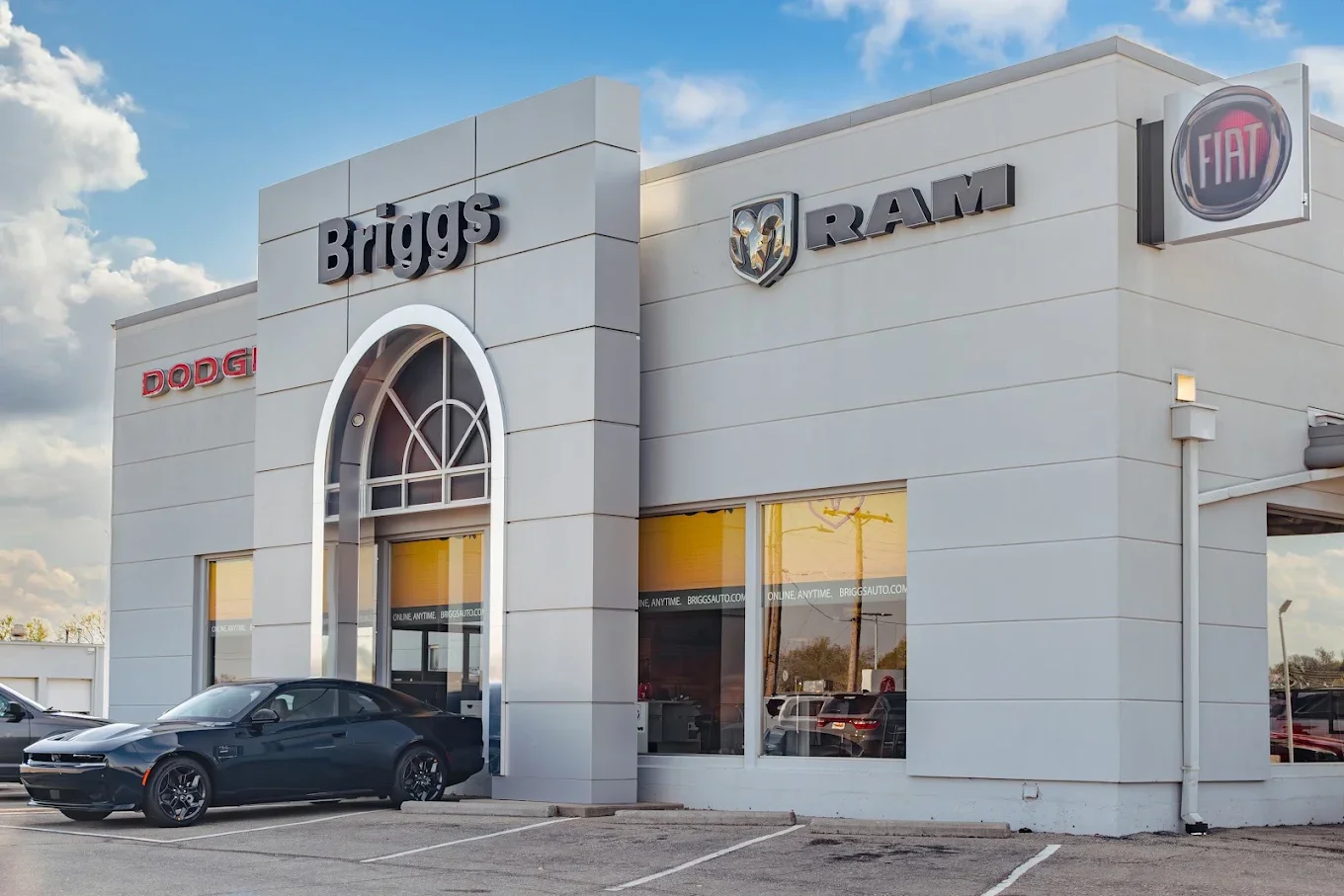 The Briggs Dodge Ram FIAT building, a pale gray showroom under a blue sky, with Briggs, Ram, Dodge and FIAT signage above an arched glass entrance and a dark car parked outside.