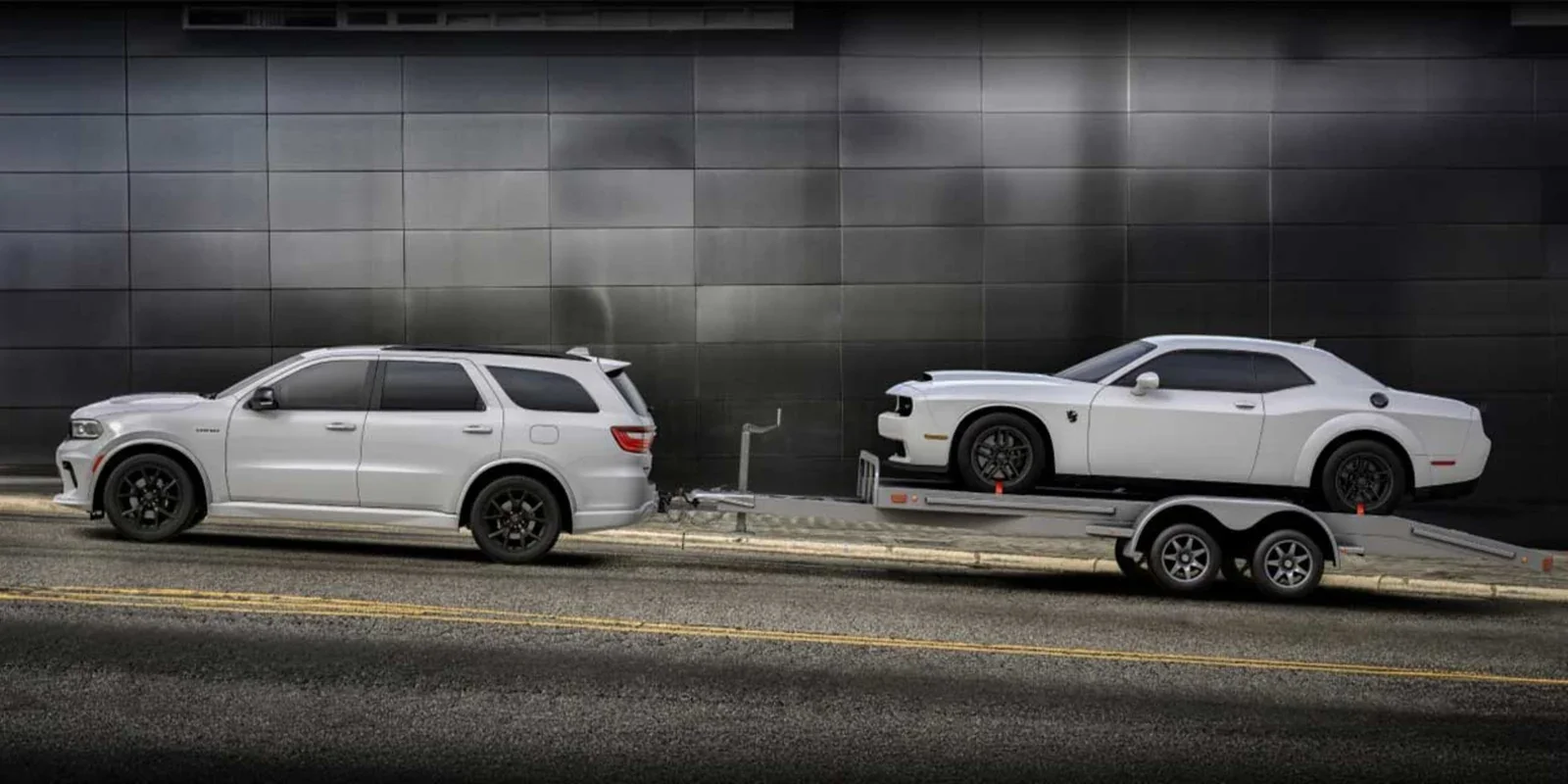 A white Dodge Durango towing a white Dodge Challenger on a car trailer, parked against a dark panelled wall.