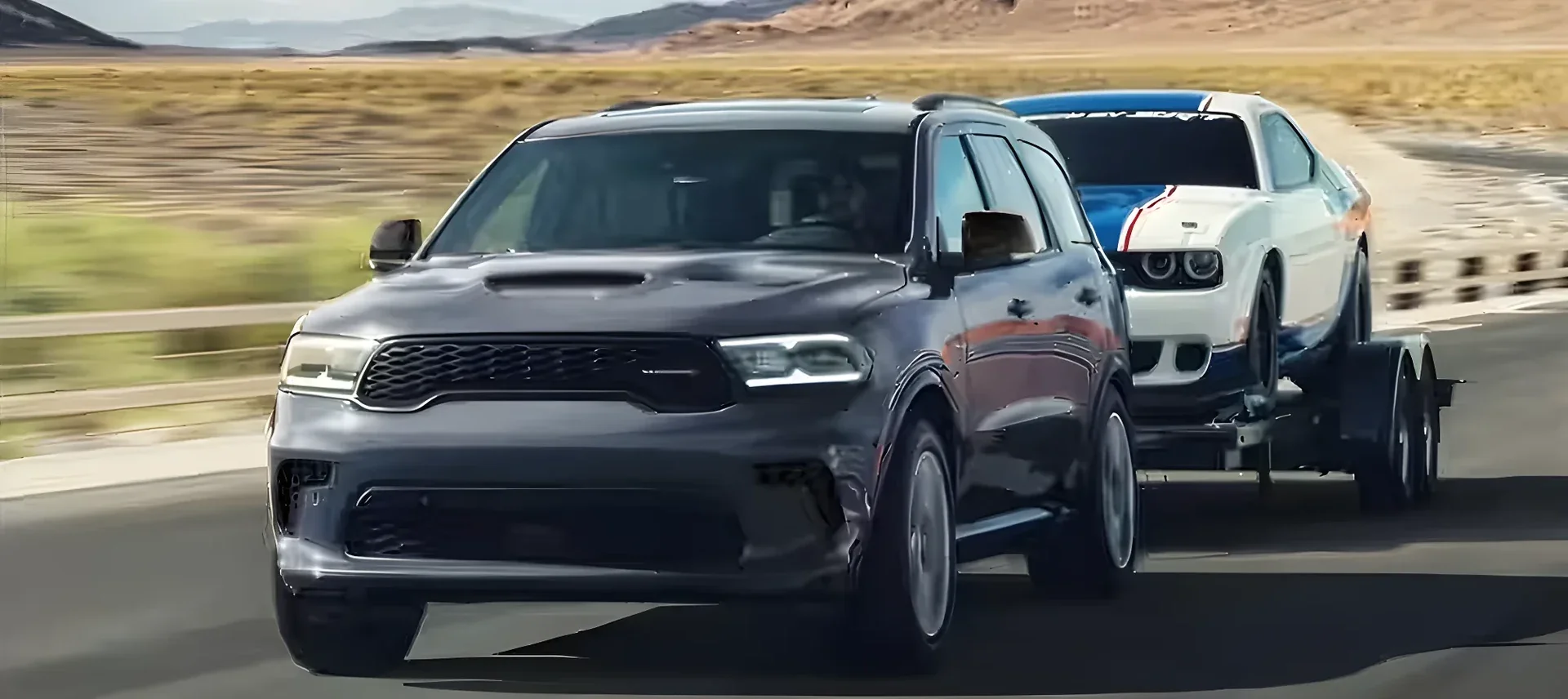 A dark Dodge Durango towing a Challenger on a car trailer down the highway.