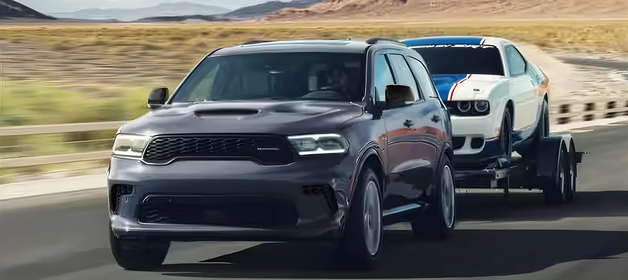 Grey Dodge Durango towing a blue race car on a trailer along a coastal road