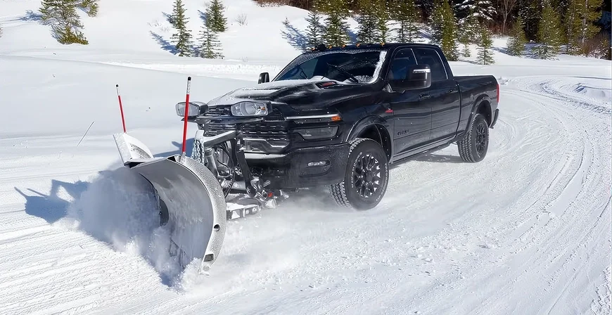 Black RAM 2500 fitted with a snowplow blade clearing a snow-covered road lined with pines