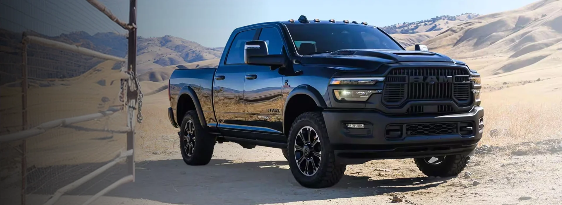 Black 2026 RAM 2500 crew cab pickup parked on a dirt track beside a wire ranch gate with dry golden hills behind it