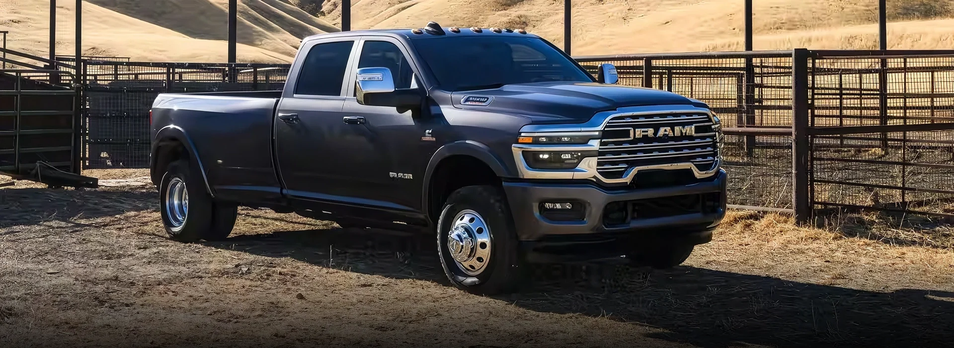Dark gray dually 2026 RAM 3500 crew cab pickup parked in a fenced dirt corral with golden hills behind it
