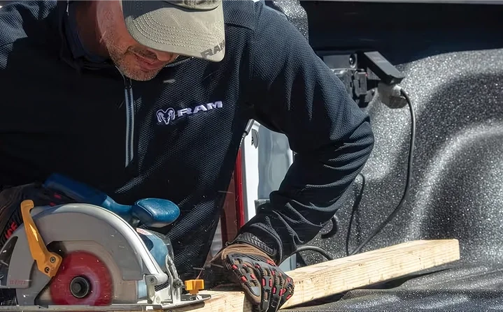 Worker in a RAM jacket cutting a board with a corded circular saw plugged into the bed of a RAM 3500