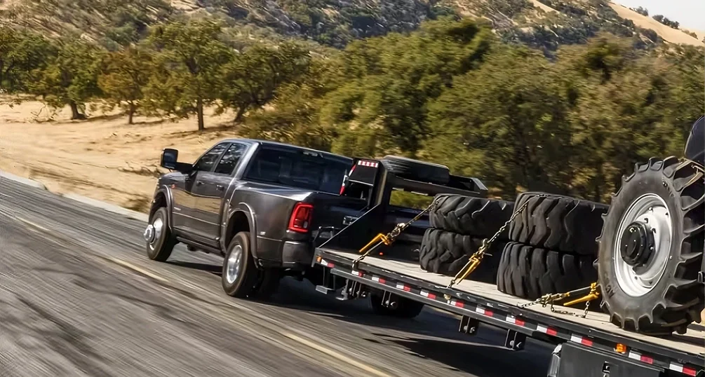 Dark gray RAM 3500 dually towing a flatbed trailer loaded with heavy equipment tires along a canyon road
