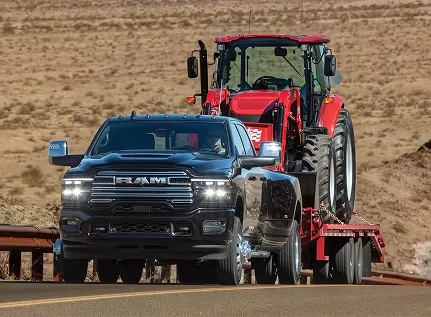 Black RAM 3500 towing a red tractor on a flatbed trailer along a desert road