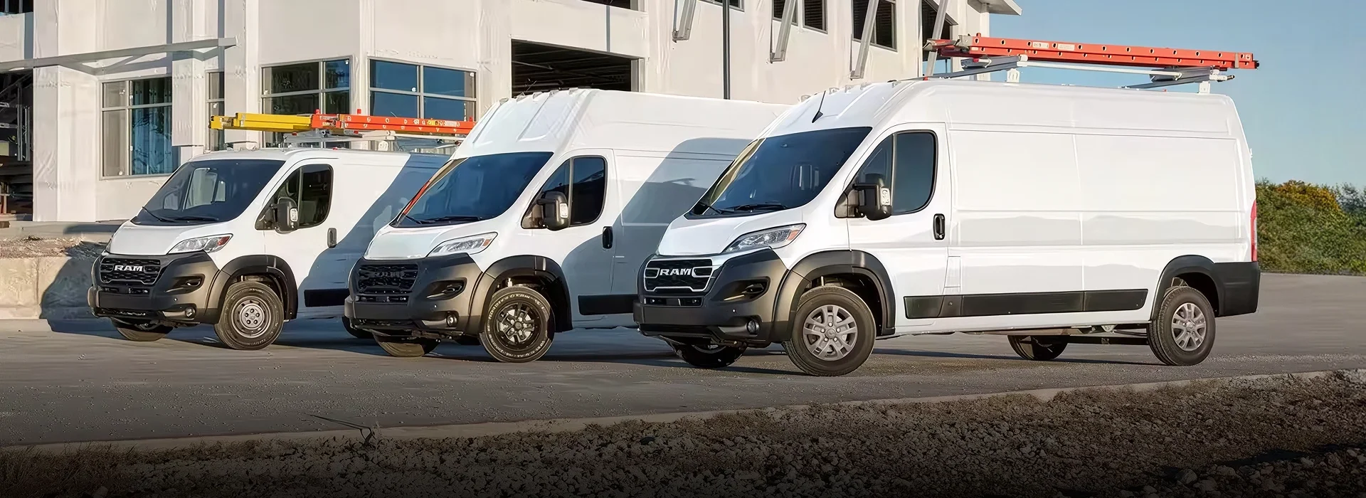 Three white RAM ProMaster cargo vans with roof ladder racks parked in a row outside a modern building at dusk