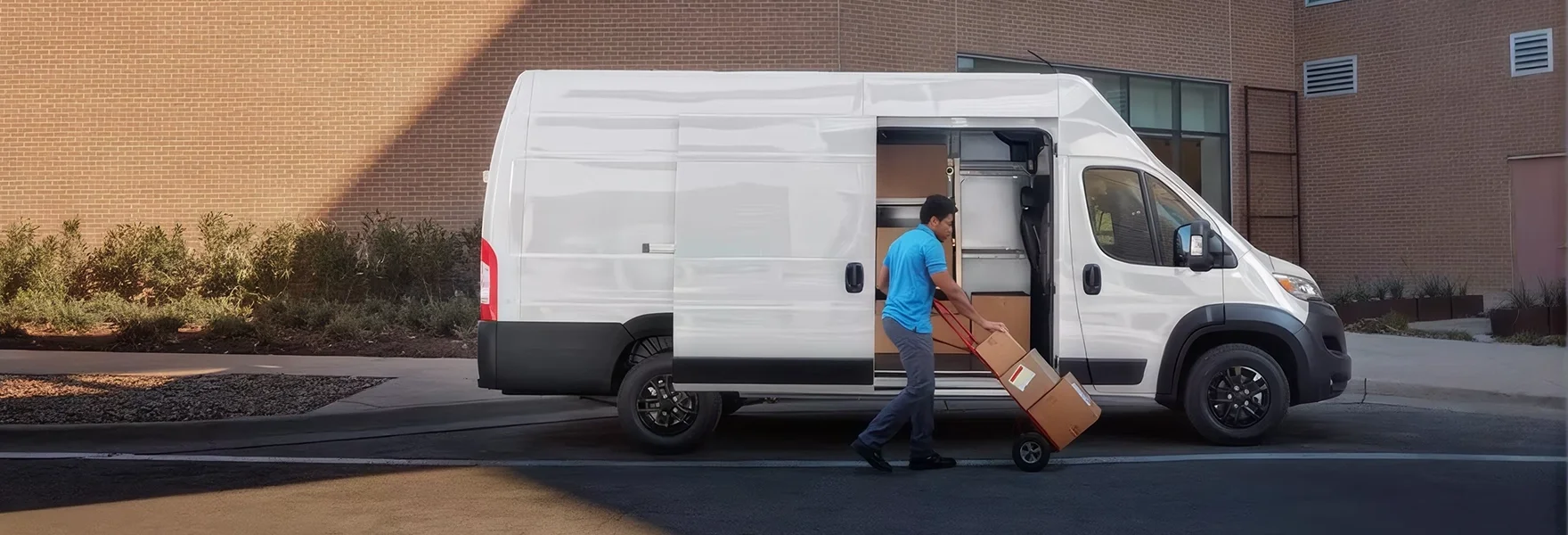Man in a blue shirt unloading boxes with a hand truck through the open side door of a white RAM ProMaster van