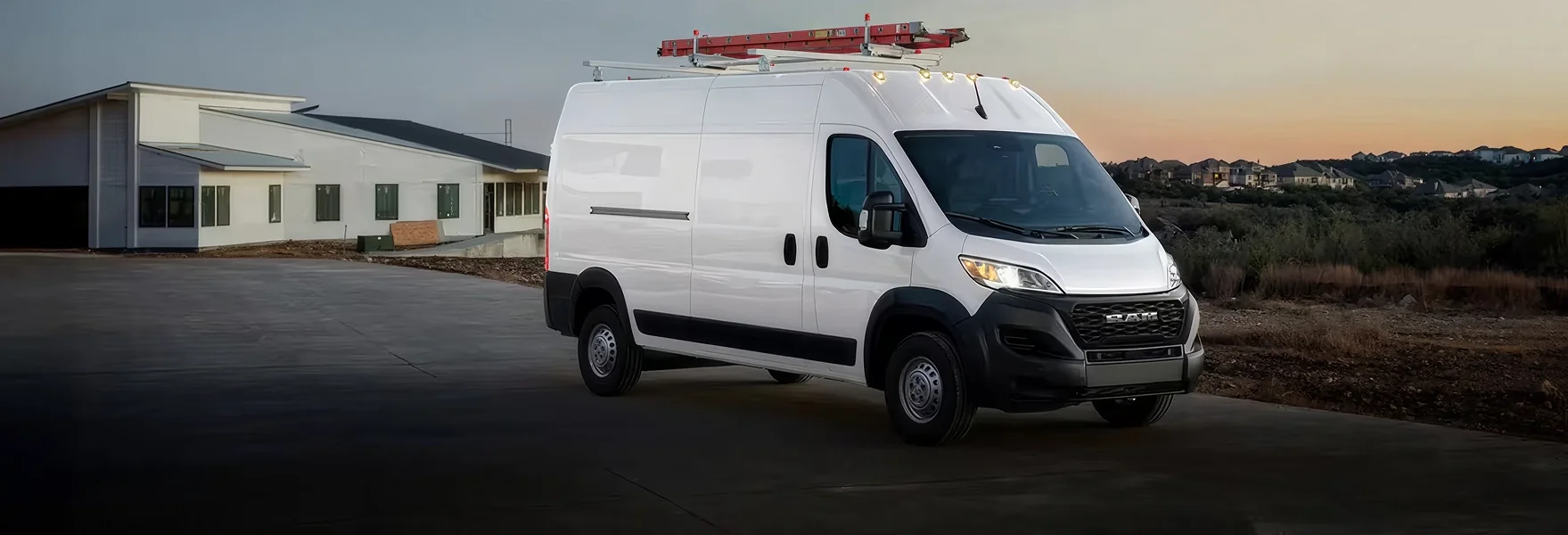 White RAM ProMaster cargo van with a loaded roof rack parked beside a building at dusk