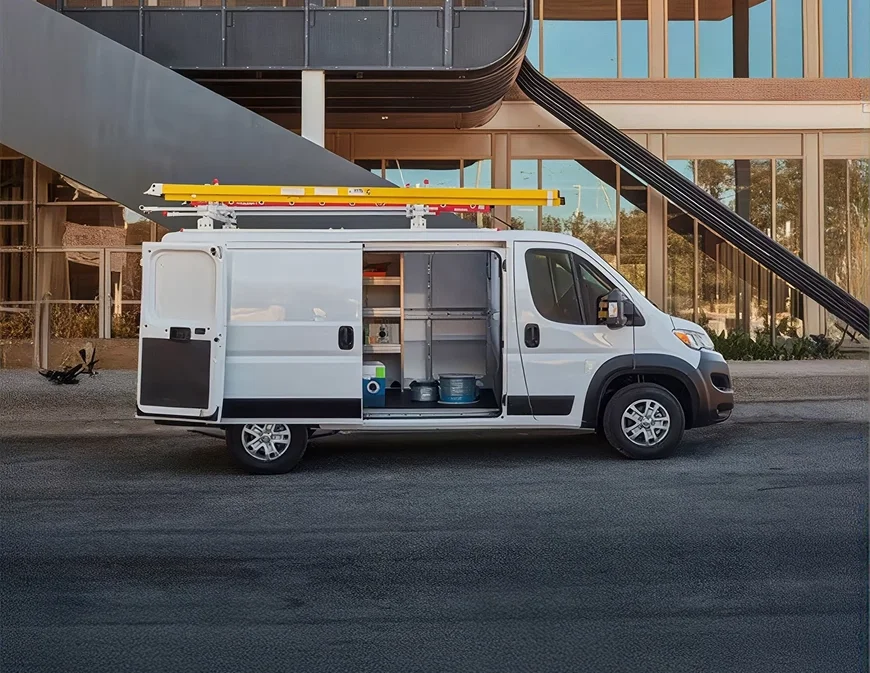 White RAM ProMaster cargo van with a ladder rack and open side door parked near a building