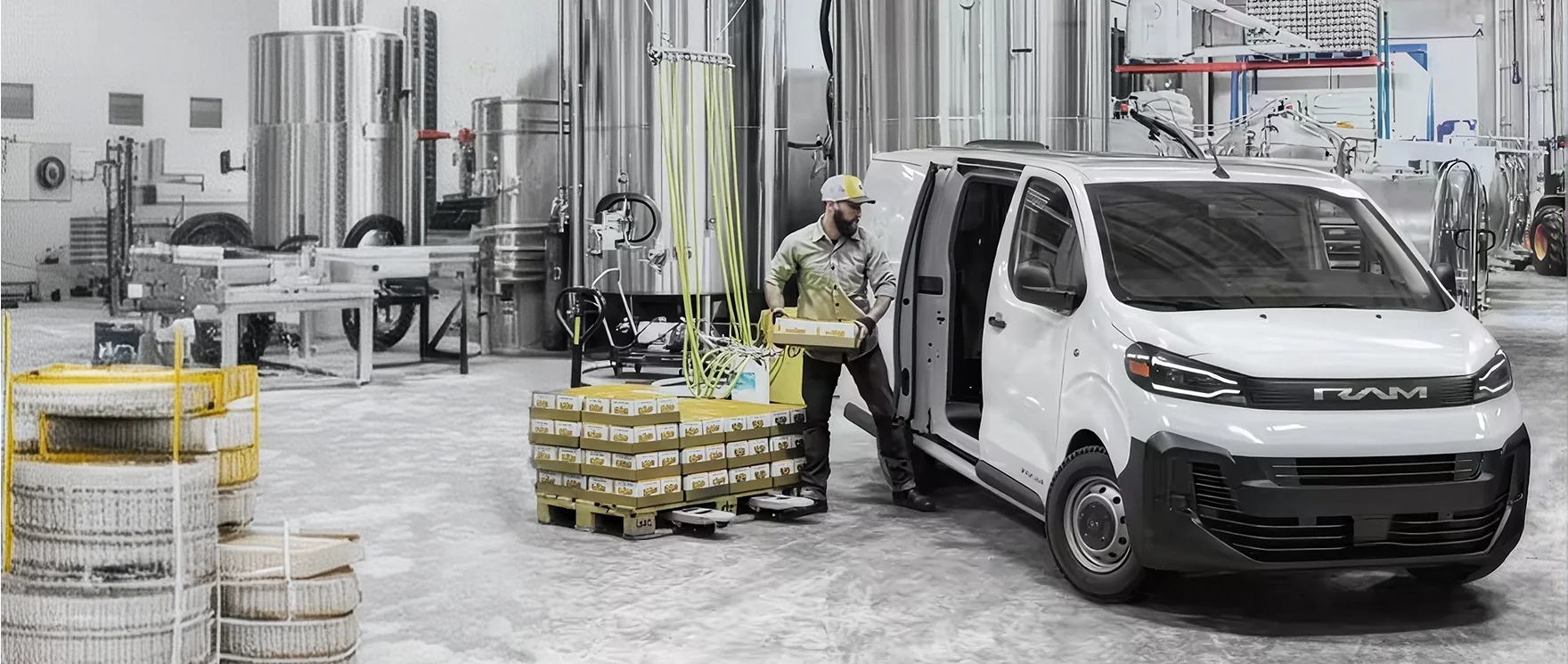 Worker in a hard hat loading a box into the open side door of a white RAM ProMaster City van beside stacked pallets in a brewery