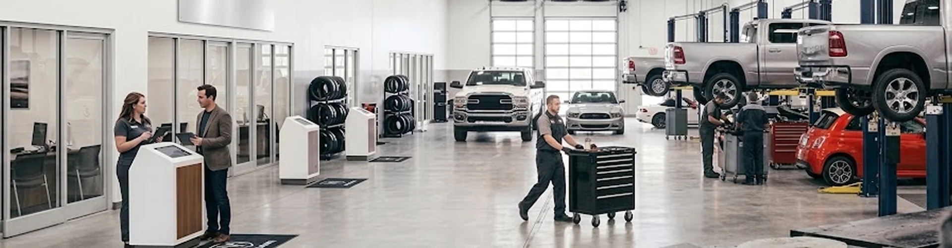 The Briggs Dodge Ram Fiat service department with a white Ram heavy duty parked between the bays, a silver Ram and a red Fiat 500 up on lifts, and an advisor helping a customer at the check-in kiosk