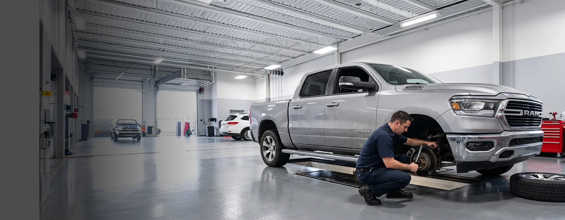A Briggs Dodge Ram Fiat technician crouched at the front hub of a silver Ram 1500 with the wheel removed, working with a wrench while the tire rests on the shop floor beside him
