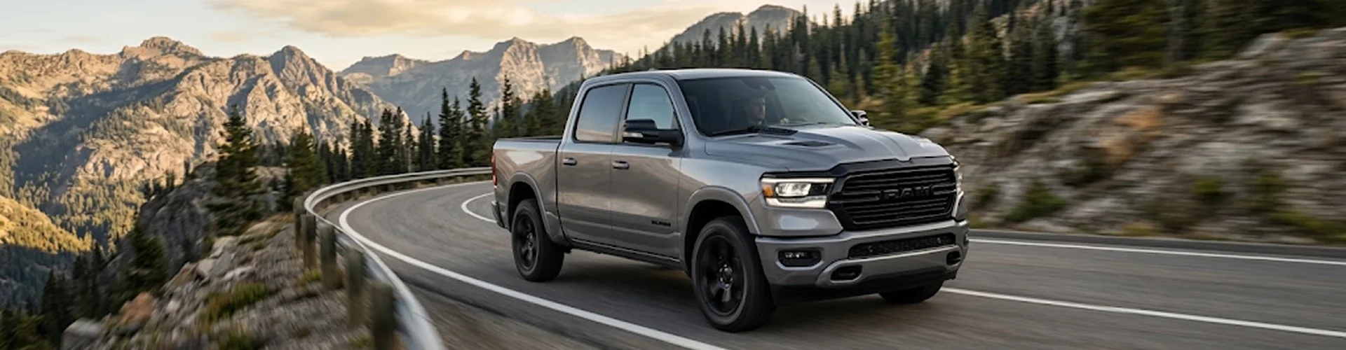 A silver Ram 1500 driving a winding mountain highway with rocky peaks and evergreens in the distance
