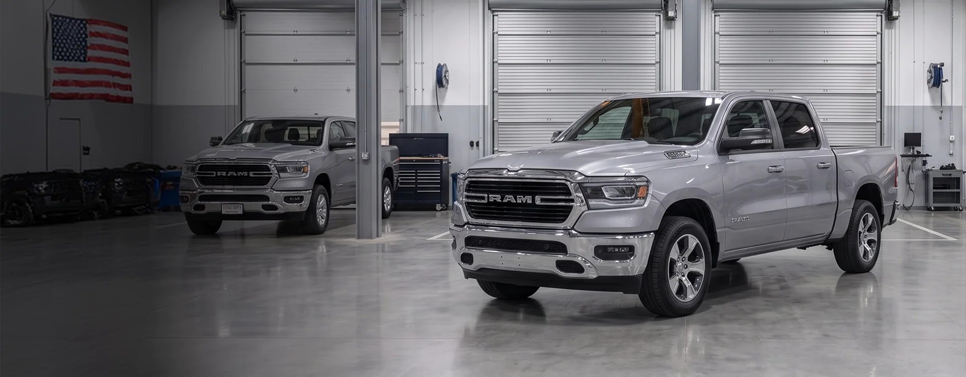 Two silver Ram 1500 pickups parked inside a Briggs Dodge Ram Fiat service bay with white roll-up doors, an American flag on the wall and a navy tool chest between them