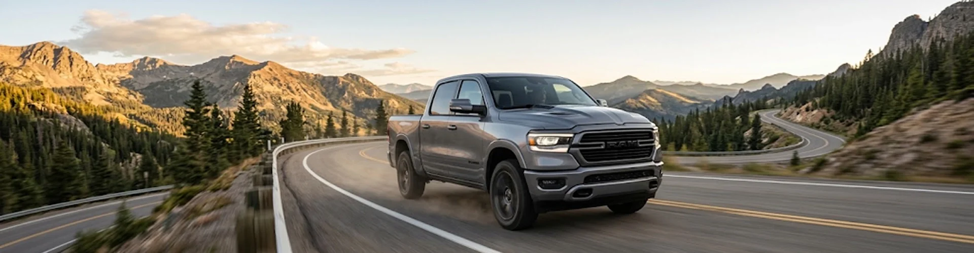 A silver Ram 1500 driving a winding mountain highway with rocky peaks and evergreens in the distance