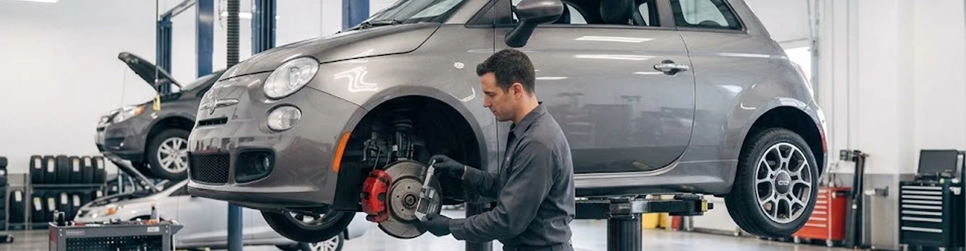 A Briggs Dodge Ram Fiat technician servicing the front brakes of a grey Fiat 500 raised on a lift, its front wheel removed to expose the rotor and red caliper