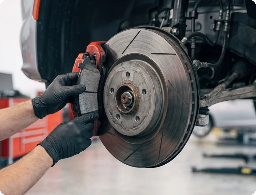 A gloved Briggs Dodge Ram Fiat technician fitting a new brake pad into the caliper of an exposed front disc brake on a raised vehicle