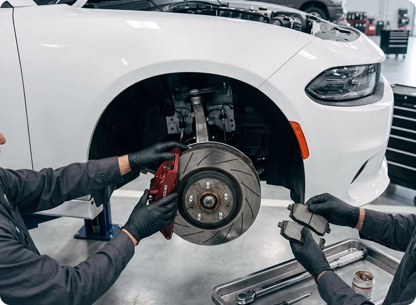 Two Briggs Dodge Ram Fiat technicians replacing the front brake pads and rotor on a white Dodge Charger, replacement pads and tools laid out on the floor beside them