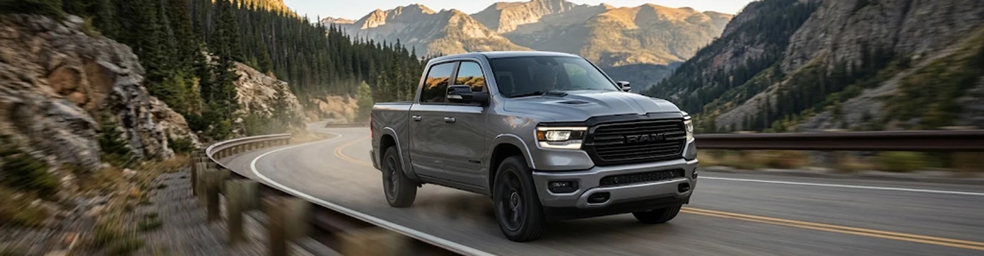 A grey Ram 1500 driving a winding mountain highway with rocky peaks and evergreens in the distance
