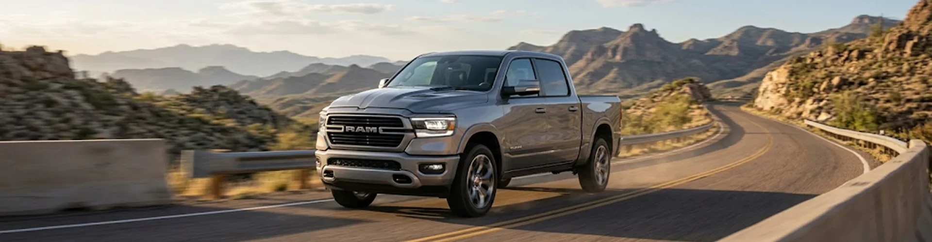 A silver Ram 1500 driving a winding mountain highway with rocky peaks in the distance
