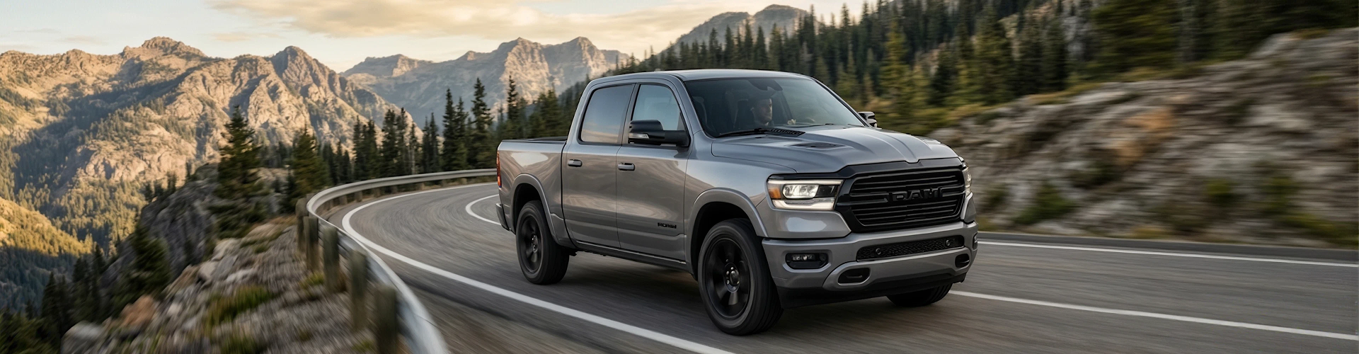 A silver Ram 1500 driving a winding mountain highway with rocky peaks and evergreens in the distance